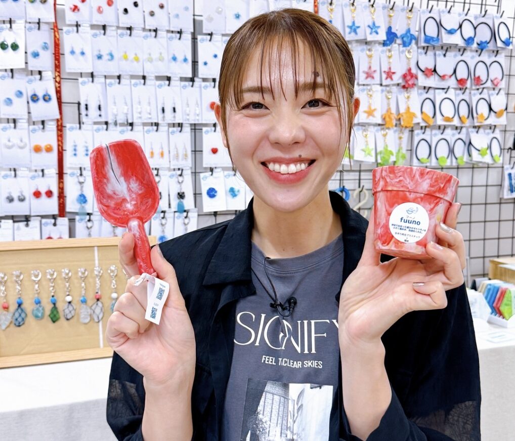 迫田さおりがショップで商品を紹介する笑顔の写真。口元のほくろが印象的なチャームポイント